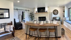 A modern kitchen with a central island, bar stools, a fireplace, and a seating area with a sofa and a chair. - Sweet Caroline Water View (Port Angeles)