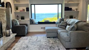 A modern living room with a large window offering a view of the sea, a fireplace, and a cozy seating area with a fluffy rug. - Sweet Caroline Water View (Port Angeles)