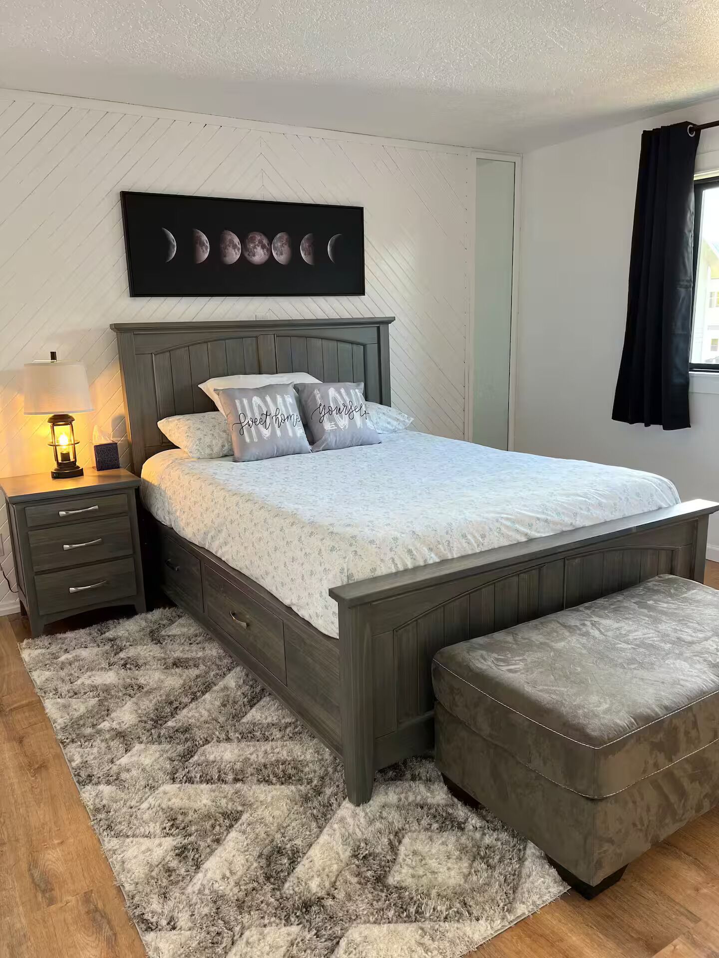 Un dormitorio con cama, mesitas de noche, un banco, una alfombra y una pared decorada con fases lunares.