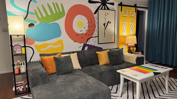 Een moderne woonkamer met een hoekbank, een salontafel en een kleurrijke abstracte muurschildering.