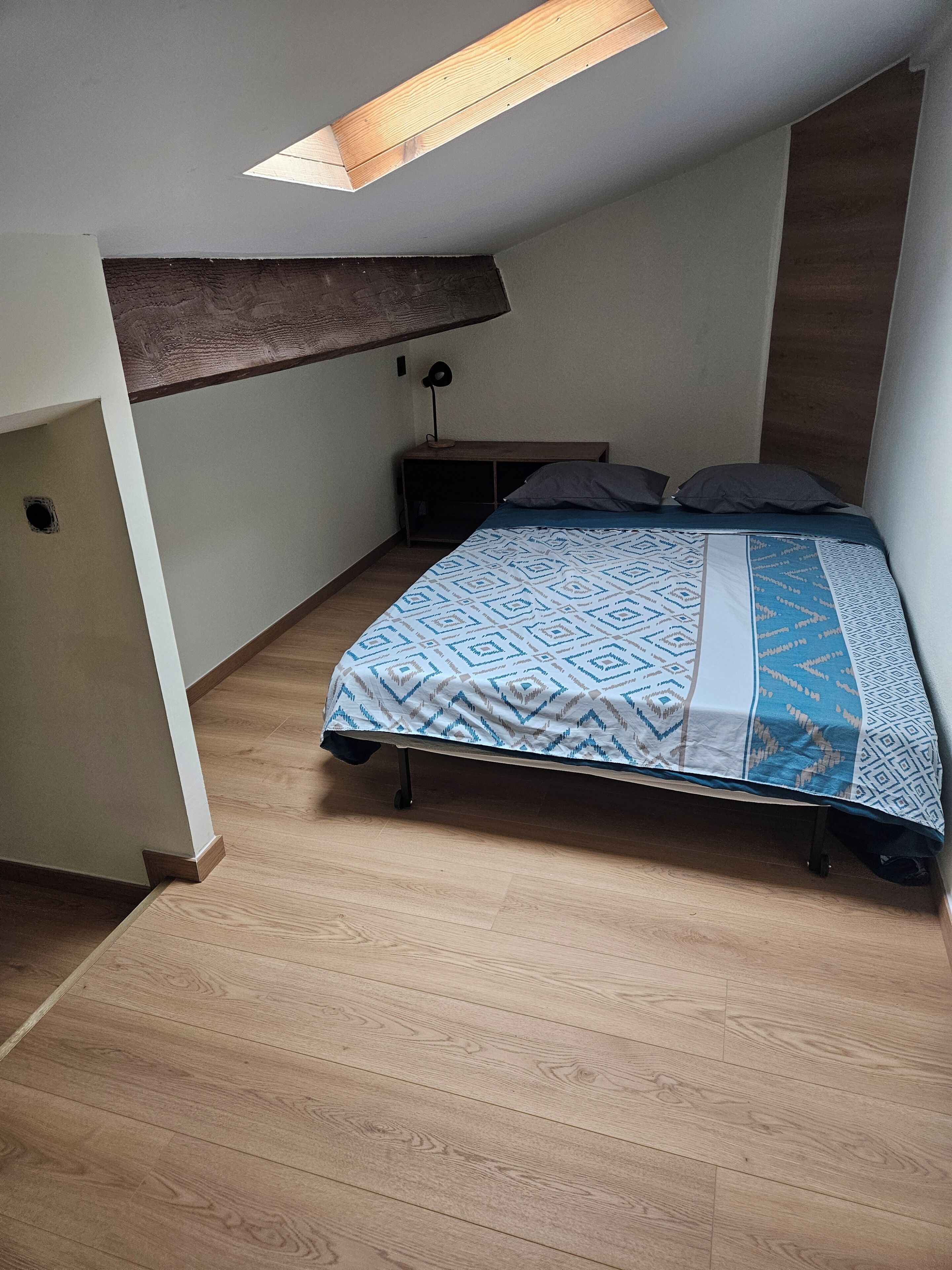 A bedroom with a bed, a nightstand, a desk, and a skylight.