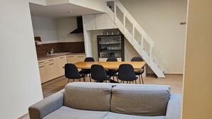 Un salon moderne avec un canapé gris, une table basse en bois et un coin repas comprenant une table et des chaises. Un escalier mène à un niveau supérieur où se trouvent une cuisine et une étagère intégrée.