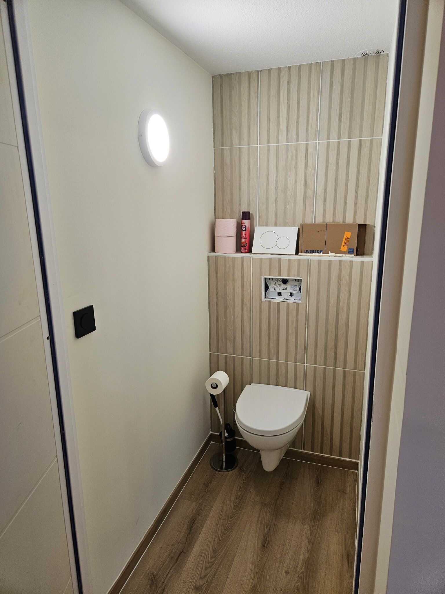 A small bathroom with a toilet, a paper towel dispenser, a soap dispenser, and a small shelf with boxes.