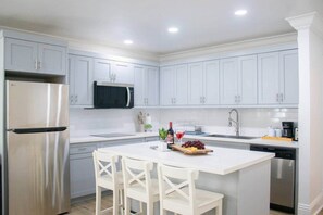 A modern kitchen with white cabinets, a stainless steel refrigerator, a microwave, and a dining area with a white table and chairs. - Tangerine Sunsets Pink Marlin (Nassau)