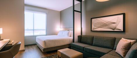 Habitación de hotel moderna con cama, sofá, escritorio y una silla. Una ventana grande ofrece luz natural y vistas.