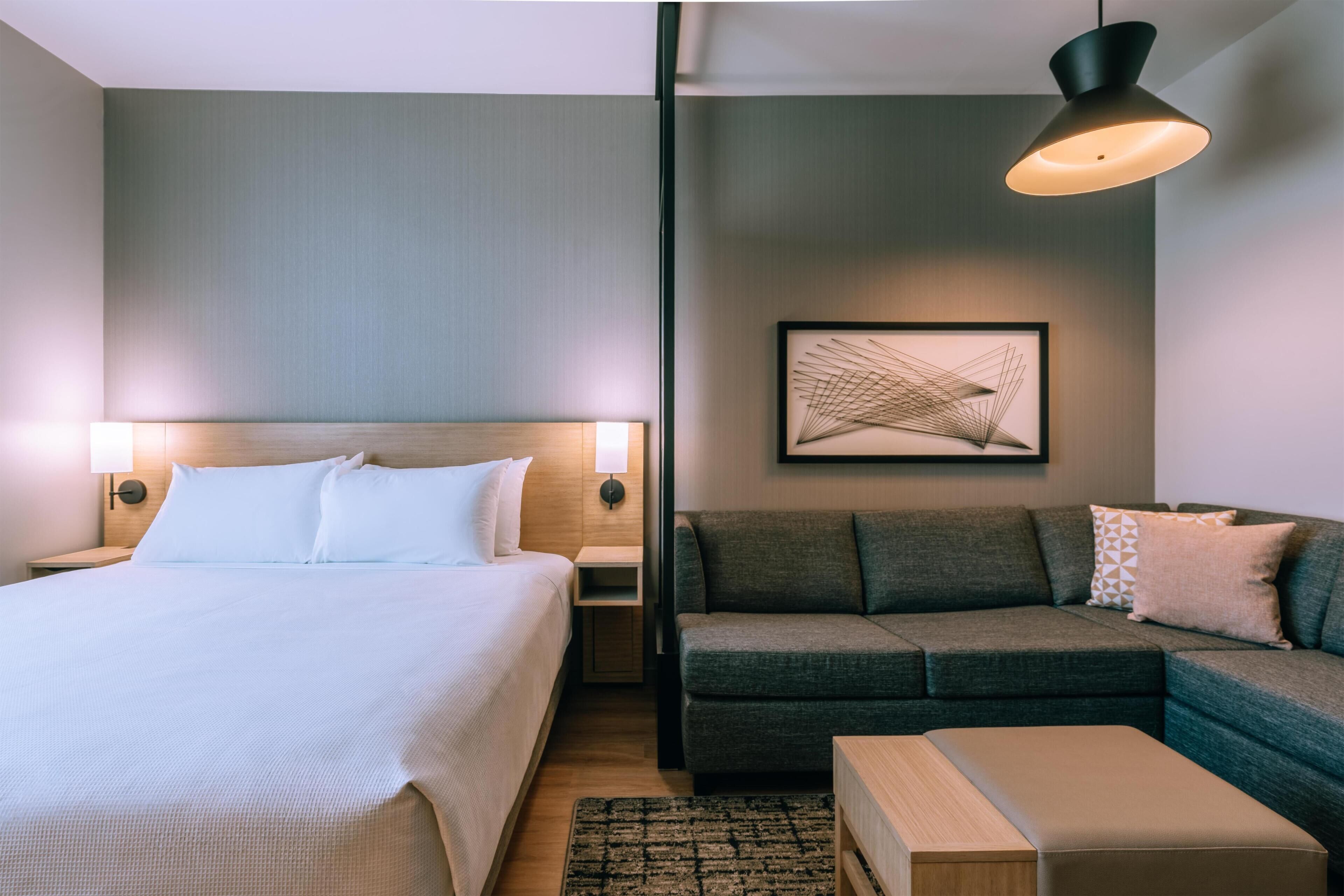 A hotel room with a bed, a sofa, a small table, and a wall-mounted artwork.