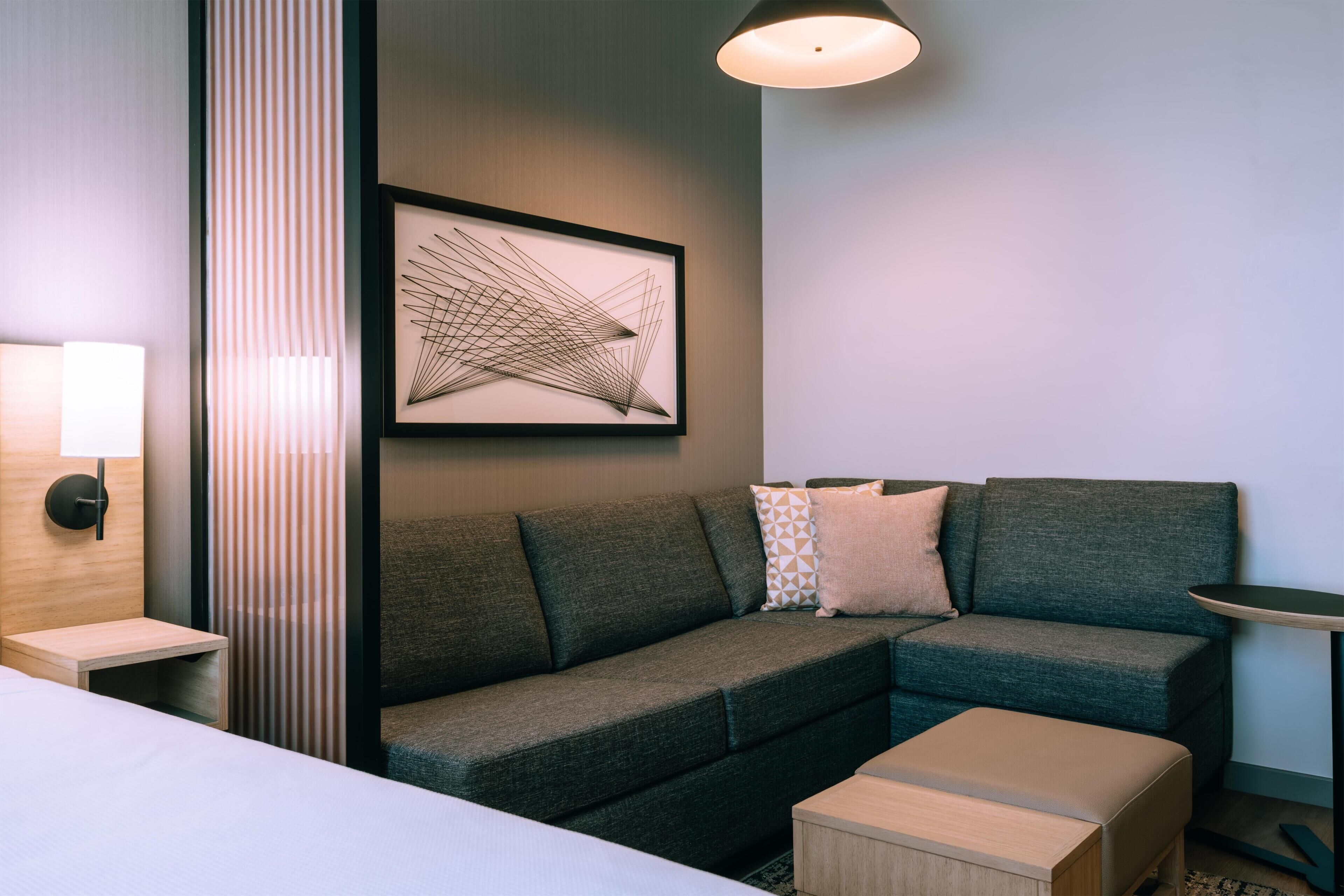 Une chambre d’hôtel moderne, équipée d’un canapé, d’une petite table, d’une lampe et d’une œuvre d’art abstraite encadrée au mur.