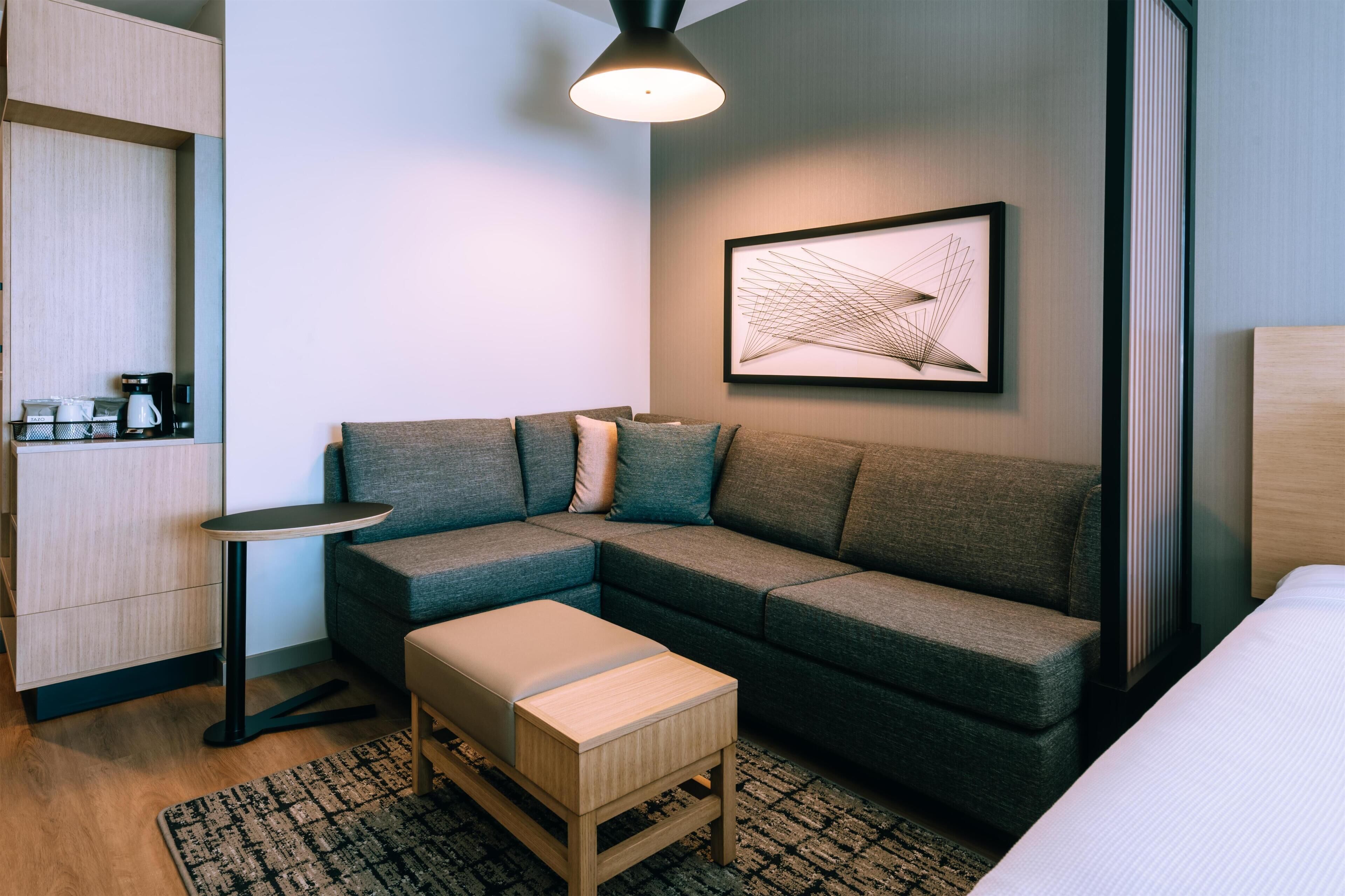 A modern hotel room with a sectional sofa, a small side table, a coffee station, and a framed abstract art piece.
