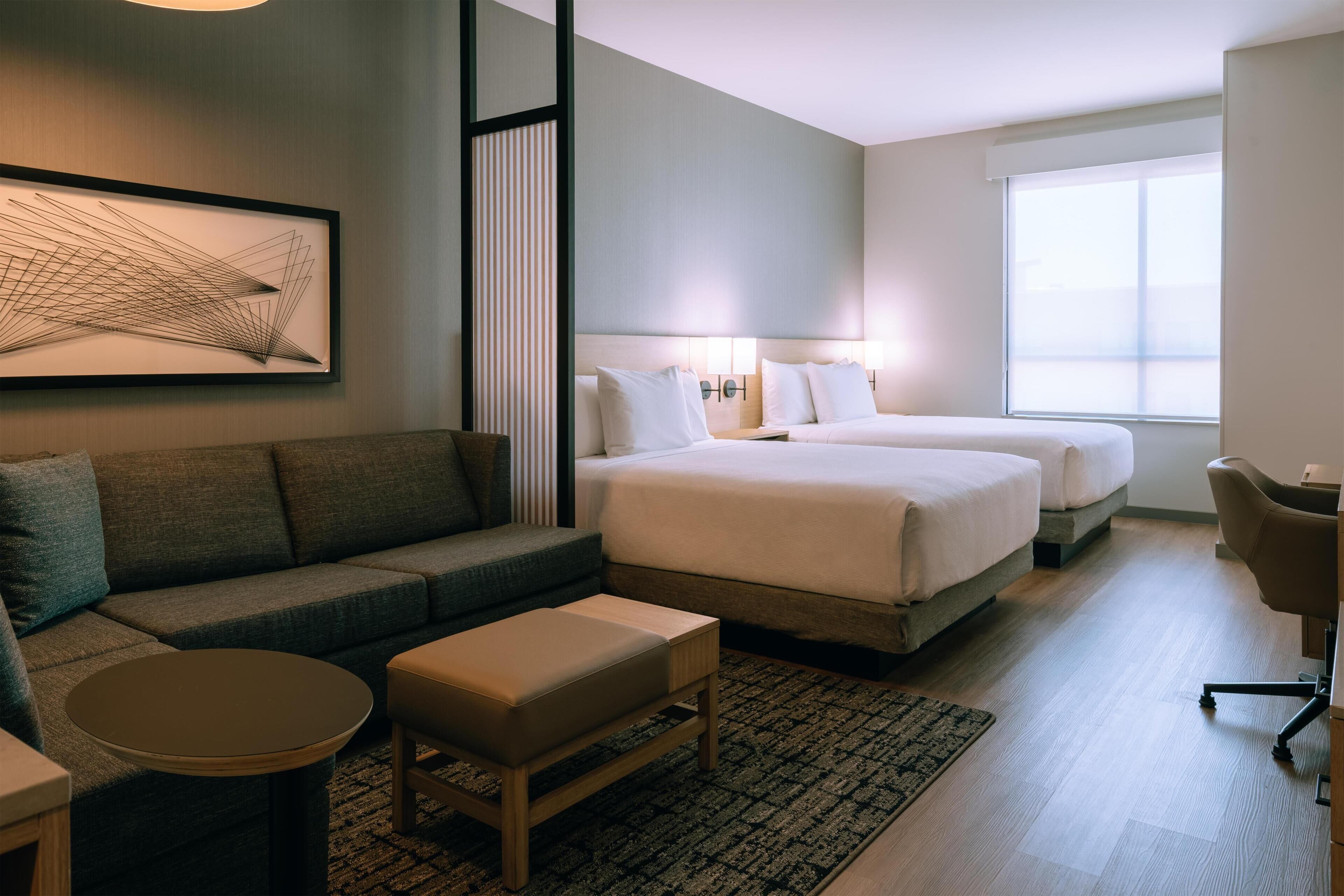 A hotel room with a sofa, two beds, a small round table, a chair, and a large window.