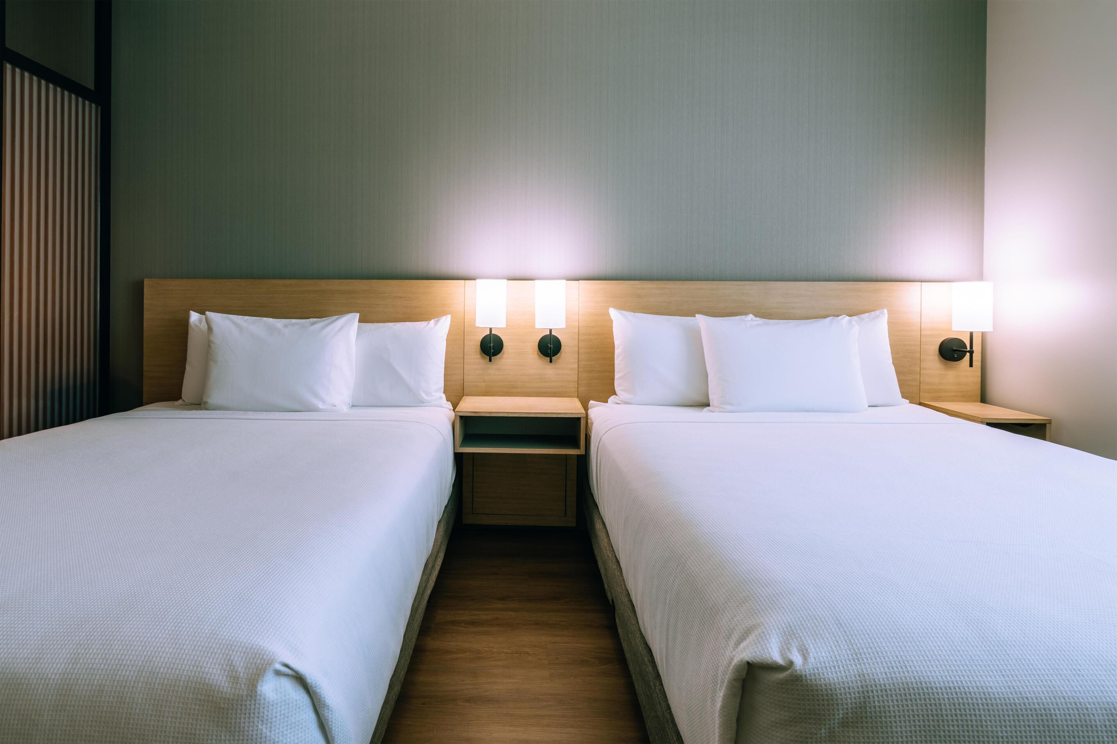 A hotel room with two single beds, each with a wooden headboard and white bedding. There are two bedside tables with lamps, and the walls are a light color.