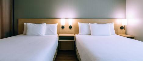A hotel room with two single beds, each with a wooden headboard and white bedding. There are two bedside tables with lamps, and the walls are a light color.