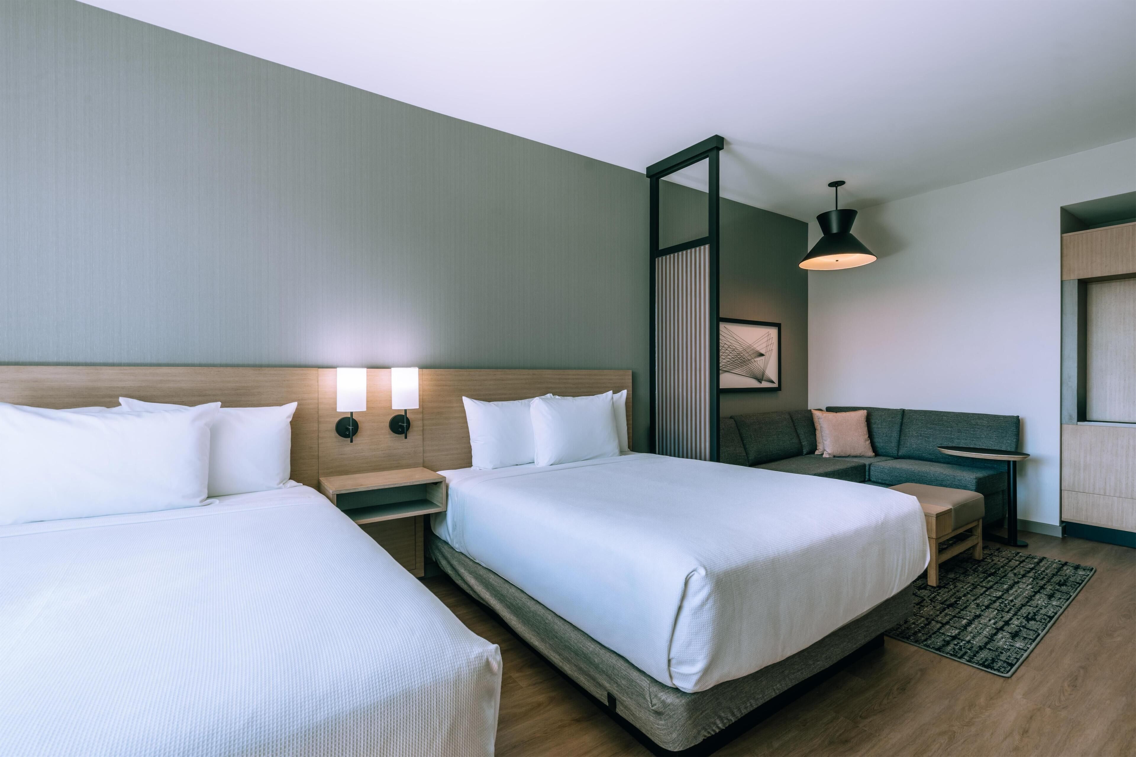 A hotel room with two beds, a nightstand, a seating area with a sofa, and a wall-mounted lamp.