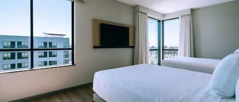 A hotel room with a large window, a bed, a flat-screen TV mounted on the wall, and a view of a building outside.