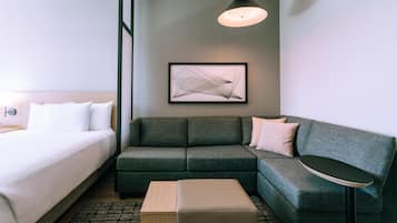 A modern hotel room with a bed, a sofa, a small table, and a framed abstract artwork on the wall.