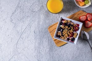 Un bol de ensalada de frutas con granola sobre una tabla de madera, un vaso de jugo de naranja y un bol de fruta mixta sobre una superficie texturizada.