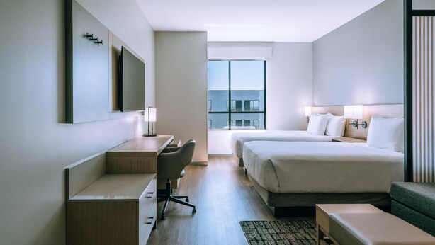 Habitación de hotel moderna con cama, escritorio, silla y vistas de la ciudad a través de amplios ventanales.