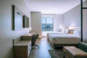 Ein modernes Hotelzimmer mit Bett, Schreibtisch, Stuhl und Blick auf die Stadt durch große Fenster.