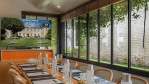 A hotel dining area with a long table set for guests, a television displaying the hotel's exterior, and large windows offering a view of the surrounding greenery.