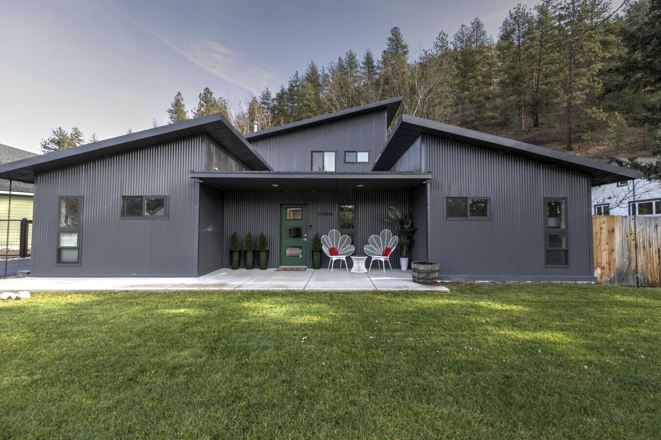 A modern two-story house with a grey exterior, a green door, and a small porch with white chairs and potted plants.