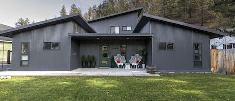 A modern two-story house with a grey exterior, a green door, and a small porch with white chairs and potted plants.