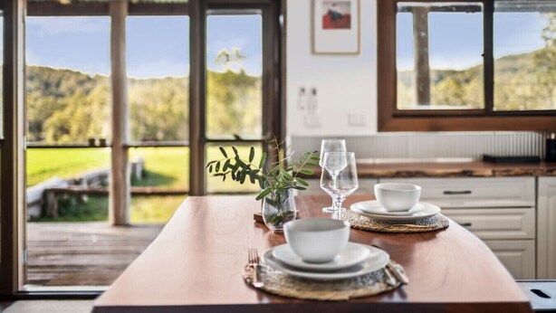 A dining table set with white dishes and glasses, a vase with greenery, and a view of a green landscape through large windows. - The Hitching Rail Hut - Relax, Unwind & Reconnect (Coramba)