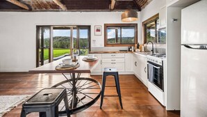 A modern kitchen with large windows, a dining area with a unique bicycle stool, and a view of the outdoors. - The Hitching Rail Hut - Relax, Unwind & Reconnect (Coramba)