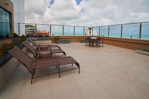 Eine Dachterrasse mit Korbsessel-Loungemöbeln und einem Tisch, mit Blick auf einen Strand und Gebäude.