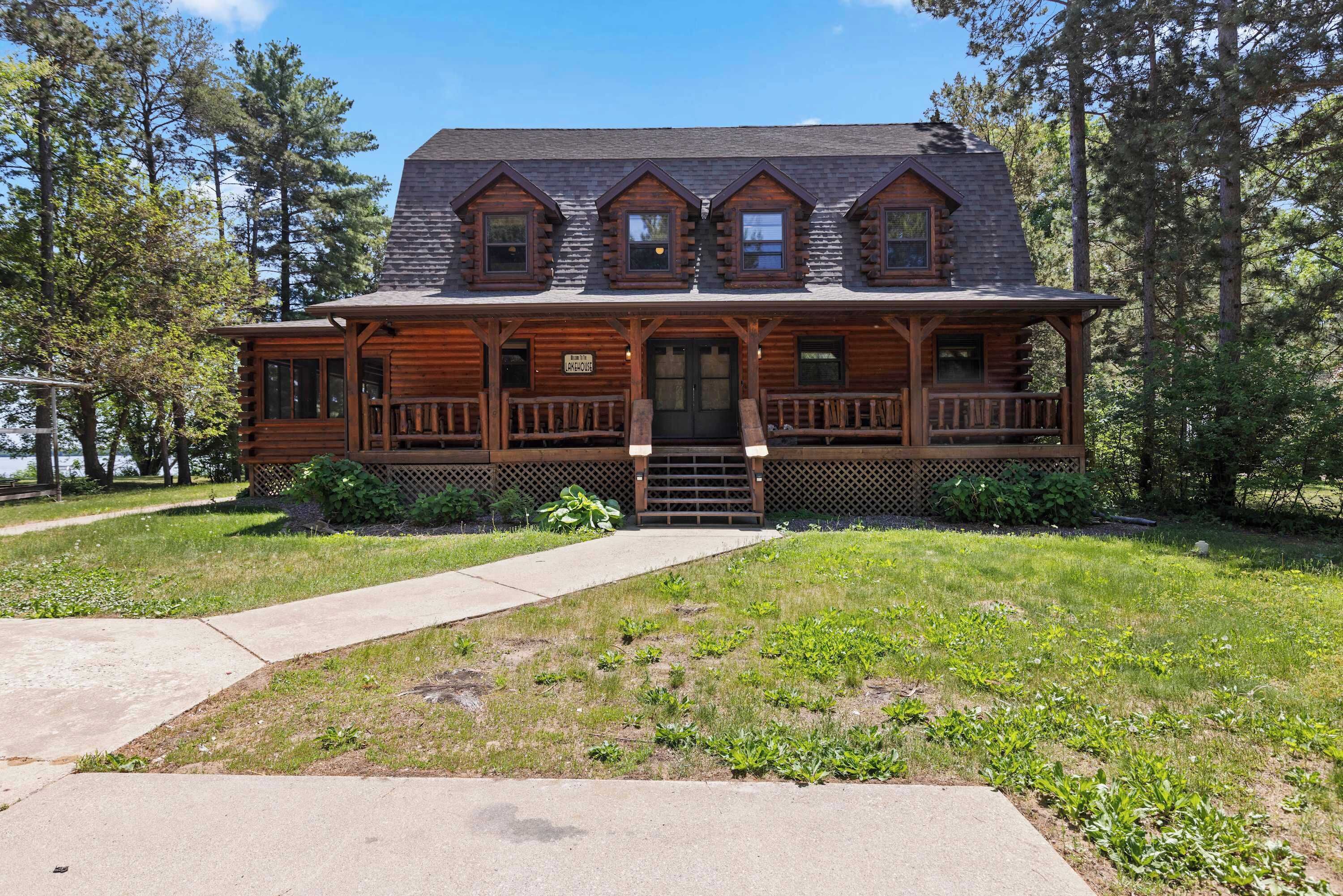 A two-story log cabin with a front porch, a central door, and a sign indicating it is a 'Lakehouse.'