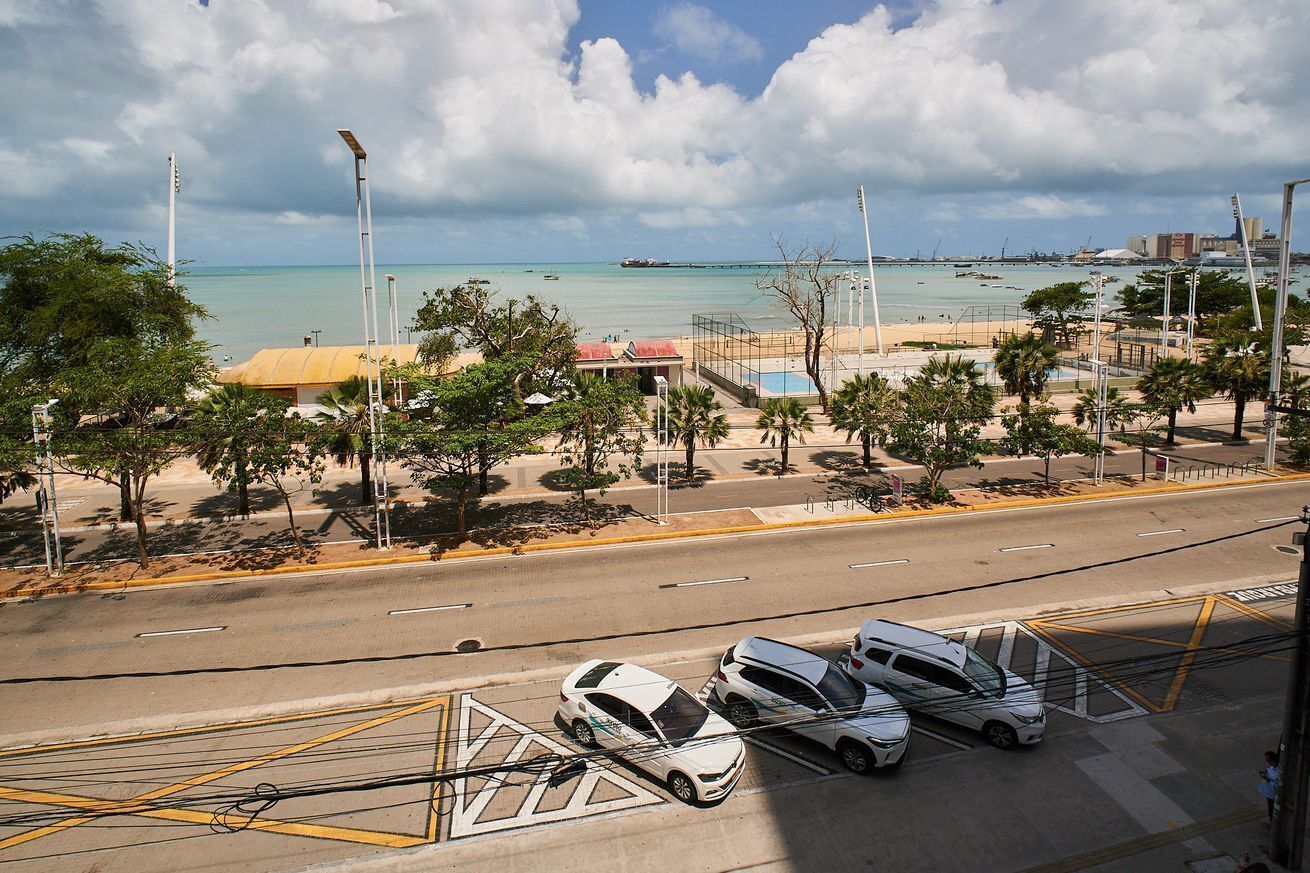 A coastal road with parked cars, a beach, and a cityscape in the background.