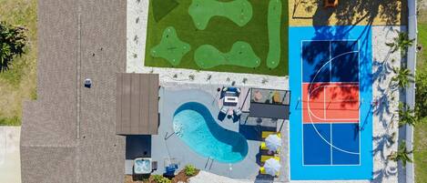 An aerial view of a backyard with a swimming pool, a basketball court, and a putting green.