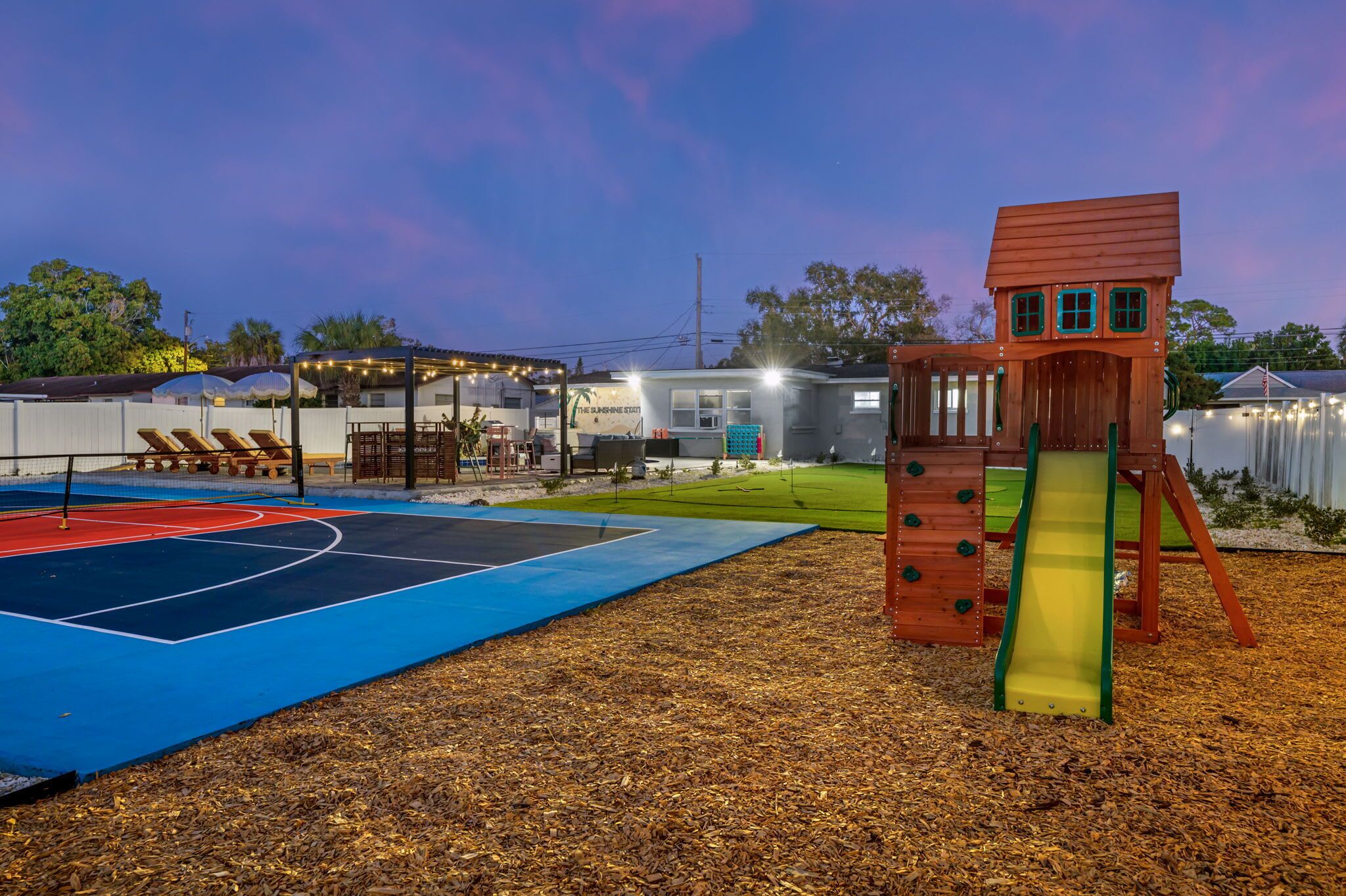 A playground with a basketball court, a slide, and a wooden playhouse.