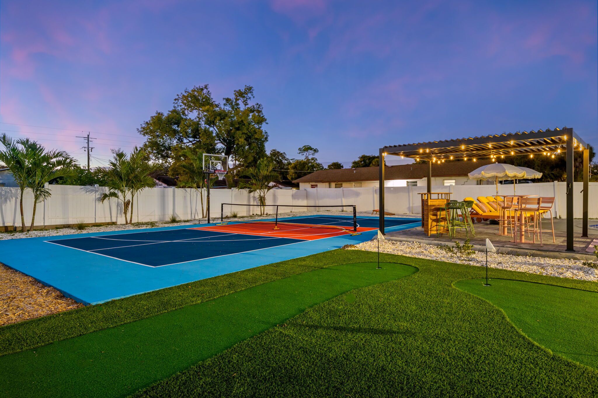 A tennis court with a net, surrounded by artificial grass and a small pool area.