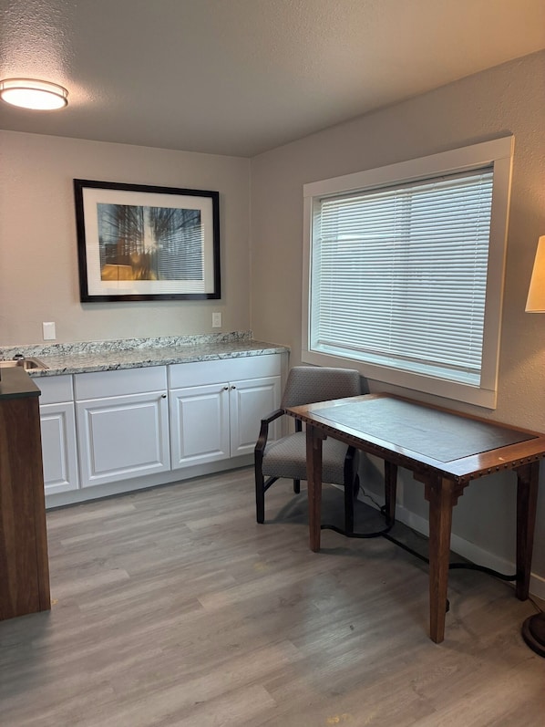 A kitchen with white cabinets, a granite countertop, a dining table with a chair, a window with blinds, and a ceiling light.