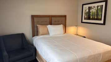 A bedroom with a bed, a chair, a bedside table with a lamp, and a framed picture on the wall.