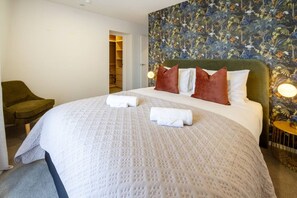 Een net opgemaakt bed met witte lakens en twee opgerolde handdoeken, een groene fauteuil en bloemenbehang.