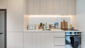 A modern kitchen with light wood cabinets, a stainless steel refrigerator, a built-in oven, and a countertop with a toaster, coffee maker, and various kitchen utensils. - Stylish & Central - City View Apt w/ Free Carpark (Christchurch)