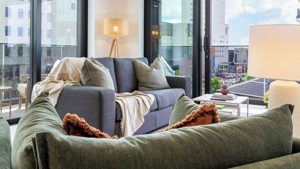A modern living room with a grey sofa, a floor lamp, and a view of a cityscape through large windows. - Stylish & Central - City View Apt w/ Free Carpark (Christchurch)