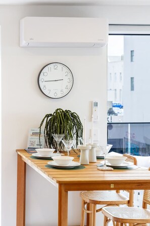 Een moderne eetruimte met een houten tafel, witte stoelen, een potplant, een wandklok en een raam met uitzicht op gebouwen.