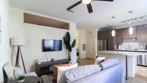 A modern living room with a gray sofa, a small round table with a plant, a TV mounted on the wall, and a ceiling fan. - Refresh Tanglewood Spacious 1BR in Uptown Houston (Houston)