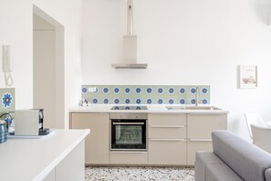 Cozinha moderna com forno embutido, bancada branca e sofá cinza na sala de estar.