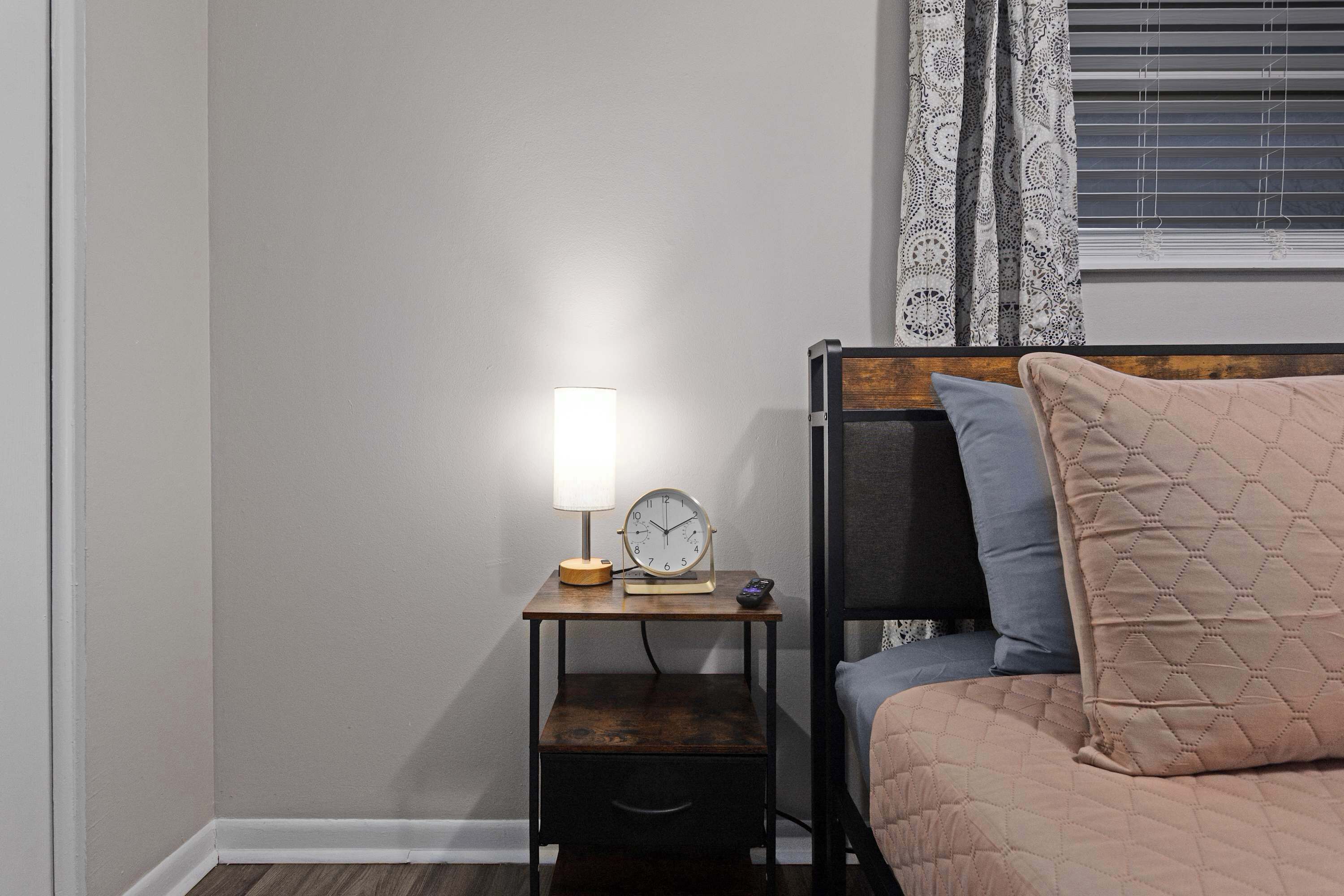 A bedroom with a bed, a bedside table with a lamp, a clock, and a small drawer.