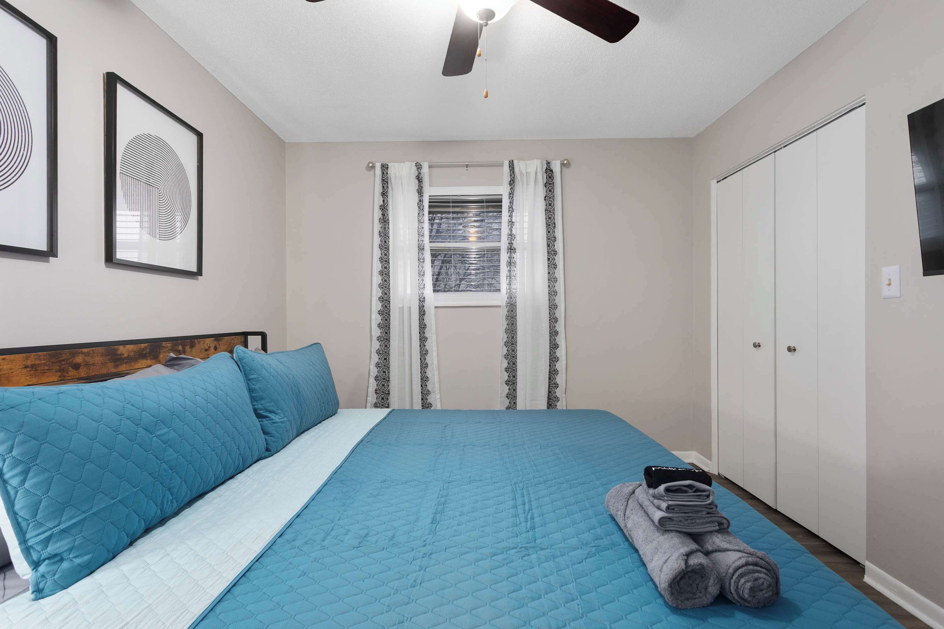 A bedroom with a bed, a ceiling fan, a window with curtains, a wardrobe, and framed artwork on the wall.
