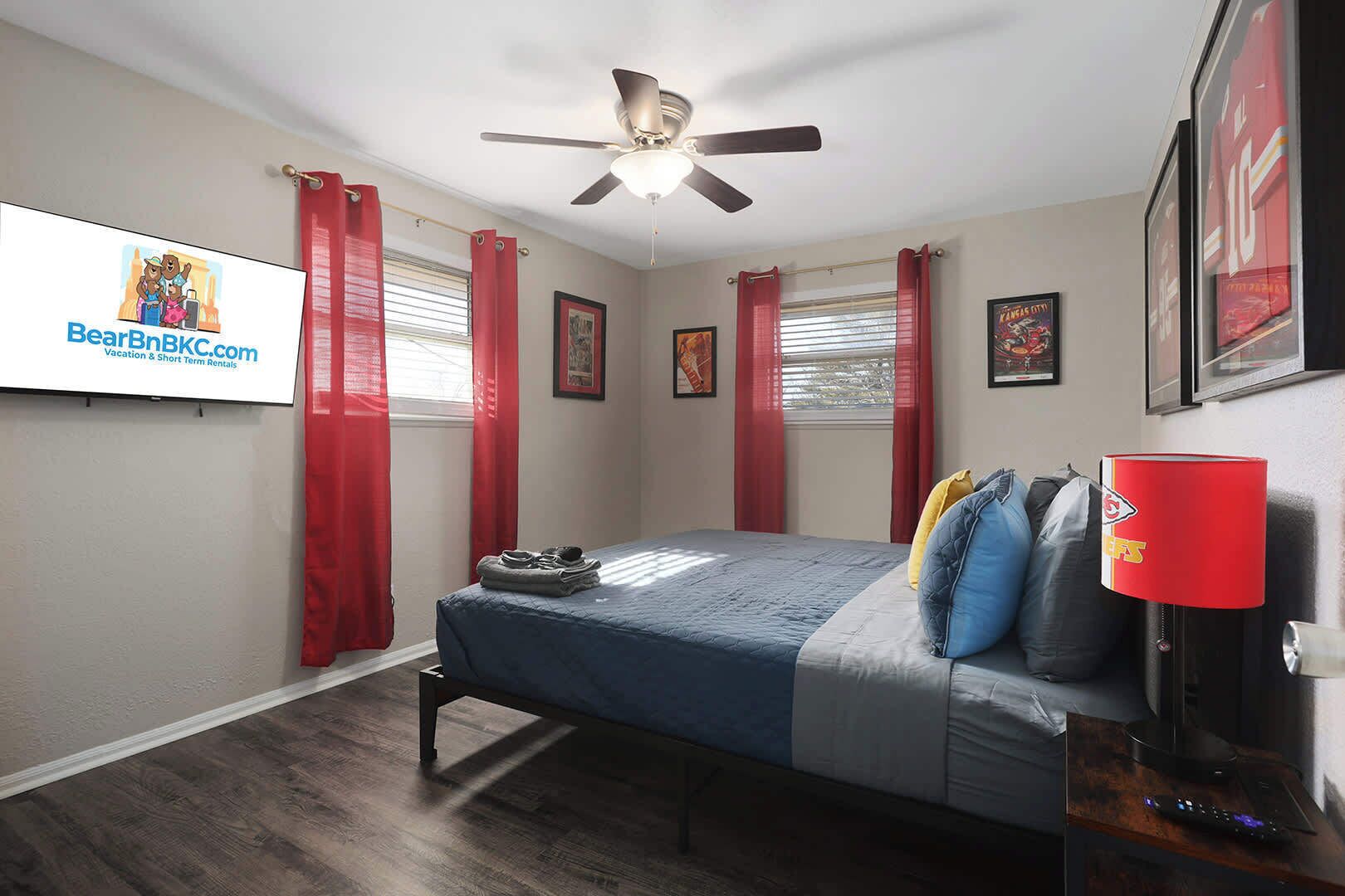 Un dormitorio con cama, ventilador de techo, cortinas rojas y un televisor mostrando BearBnB.com.