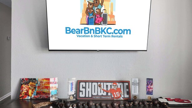 A television displaying BearBnB.com, a sign with a football player, and a row of trophies. - Chief Themed 2 BR APT_KingBeds_Near Dining (Kansas City)