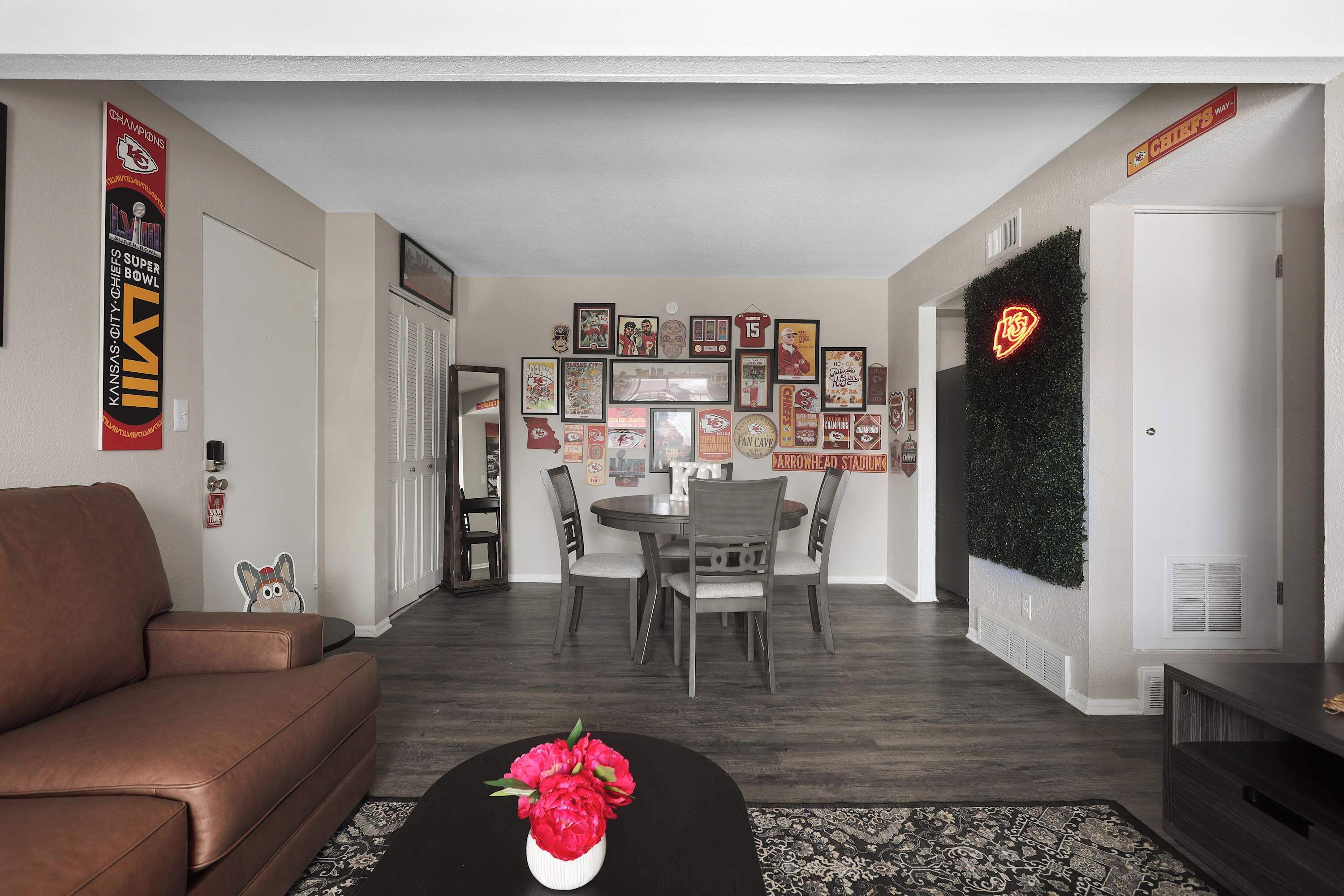 A living room with a brown sofa, a coffee table with flowers, a dining area with a table and chairs, and a wall decorated with sports memorabilia and a neon sign.