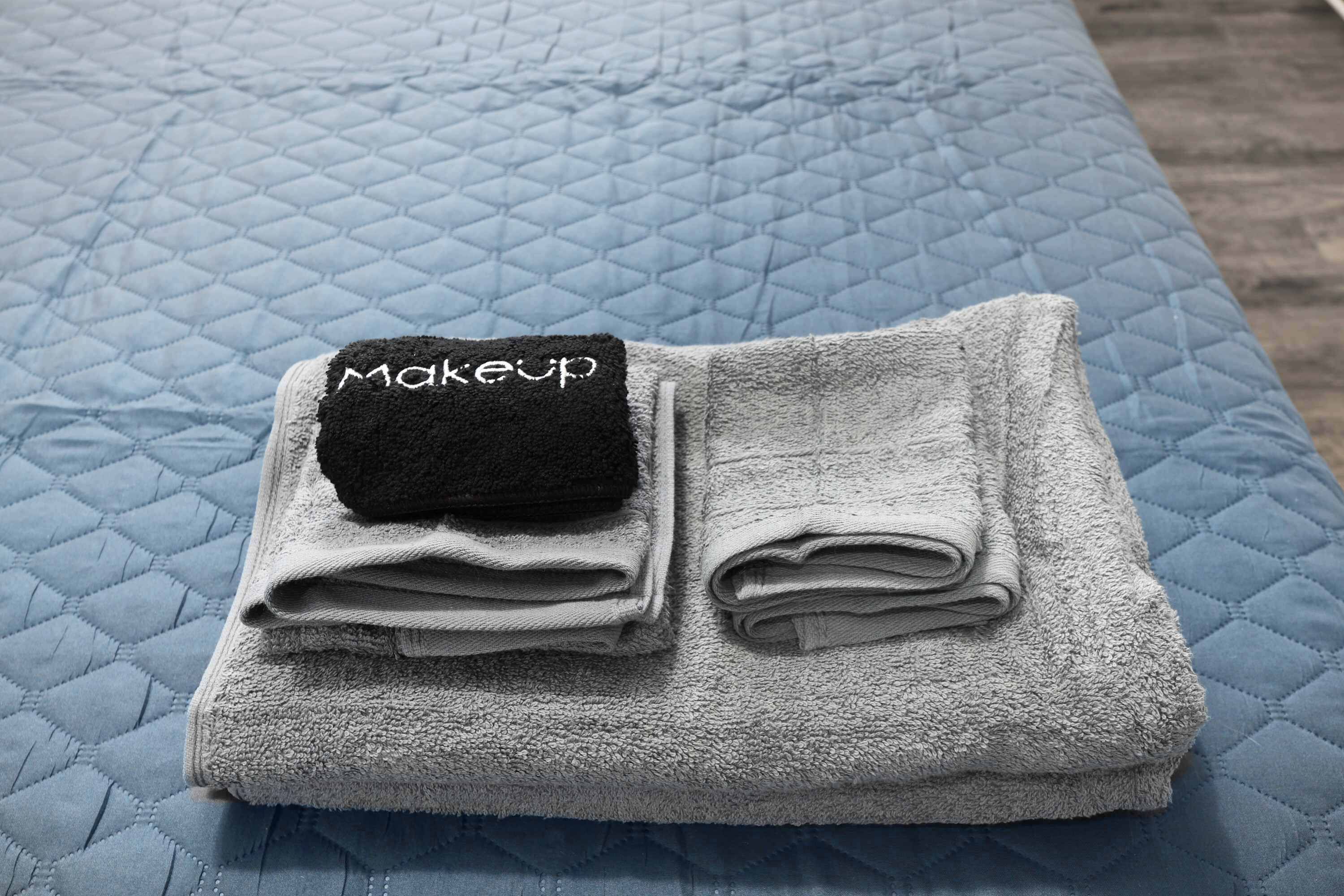 A bed with a quilted blue cover and a folded grey towel with a black tag labeled "makeup".