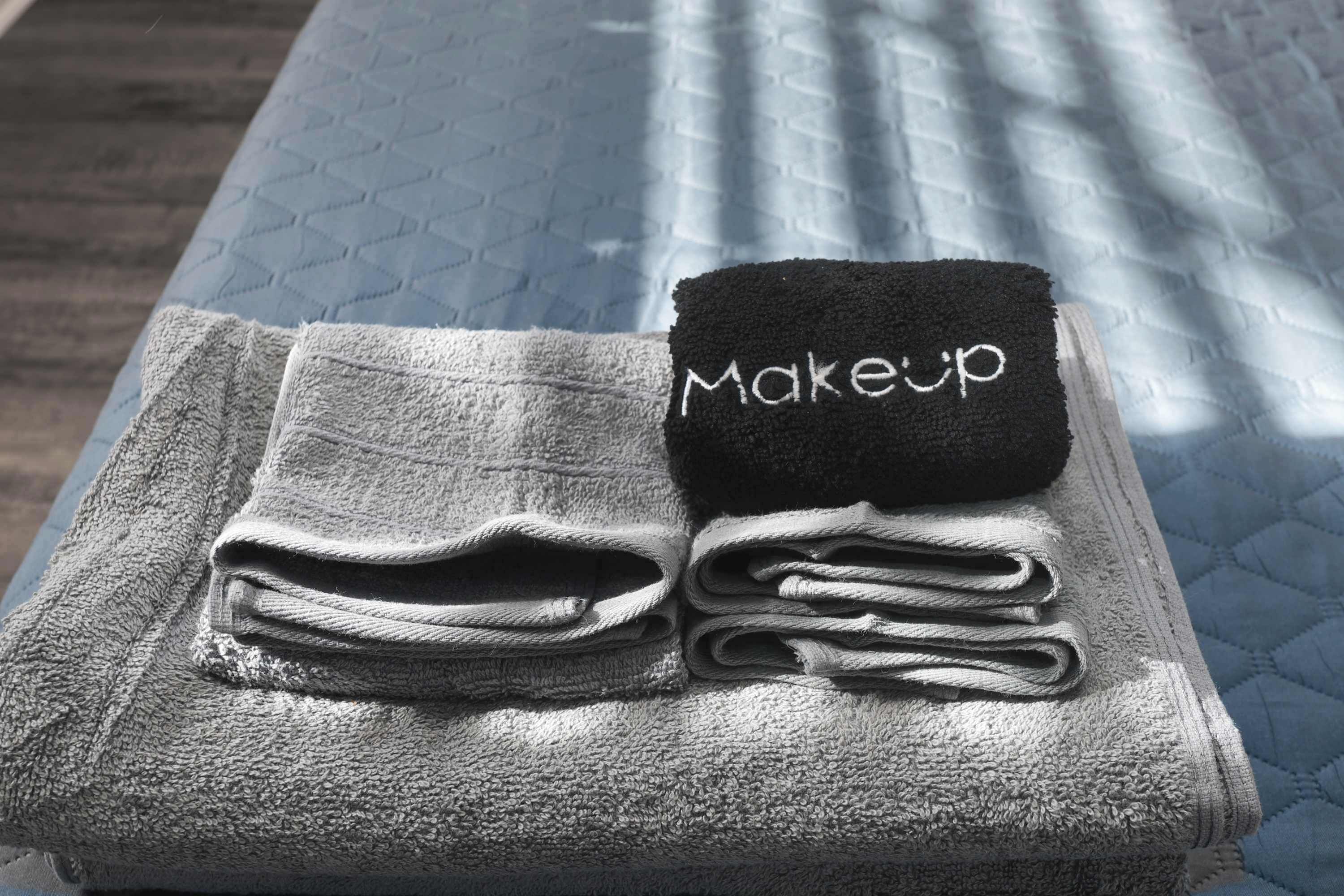 A neatly folded towel stack with a black towel labeled "Makeup" on a bed with a patterned bedspread.