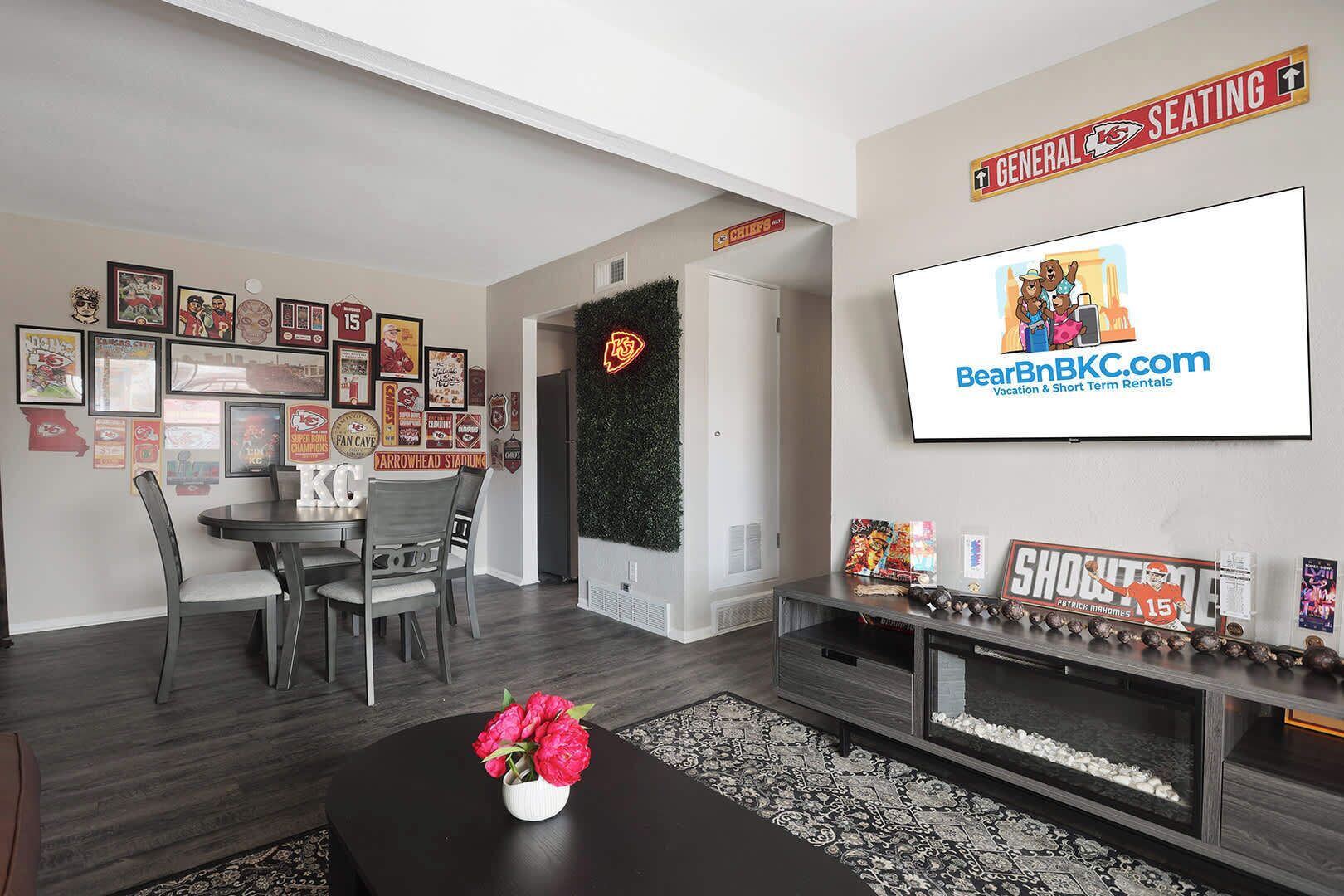 A modern living room with a dining area, decorated with sports memorabilia and a large TV screen advertising BearBnB.com.