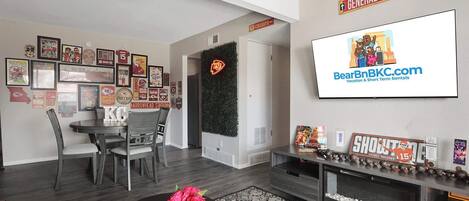 A modern living room with a dining area, decorated with sports memorabilia and a large TV screen advertising BearBnB.com.