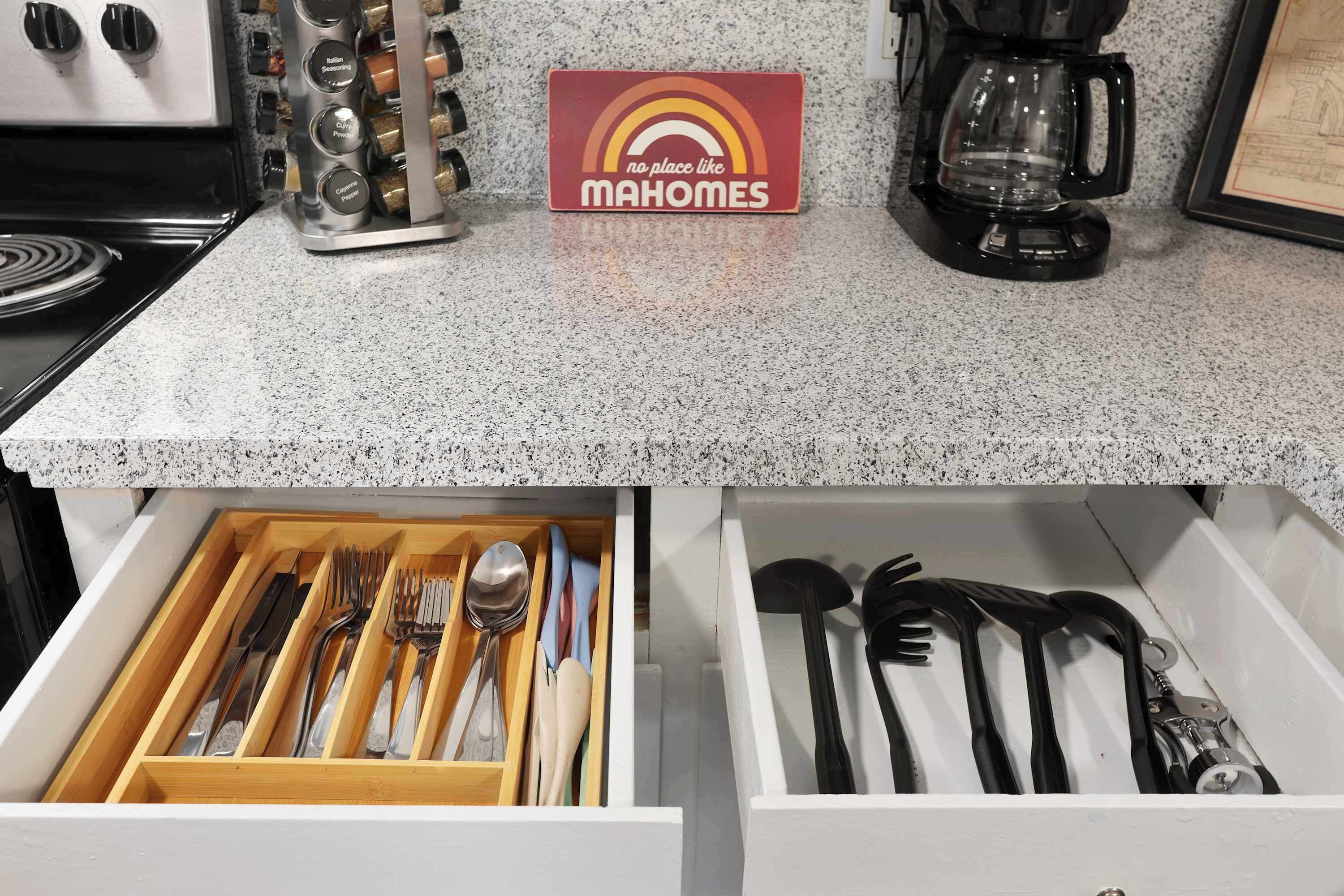 A kitchen counter with a coffee maker, a spice rack, and a sign that reads "no place like MAHOMES." Below, a drawer contains utensils and a spoon.